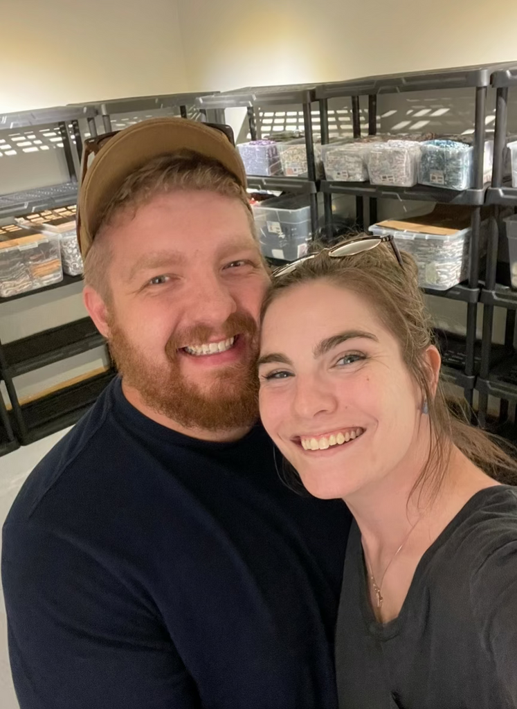 Two people posing for a selfie in an indoor storage area with shelves and containers.